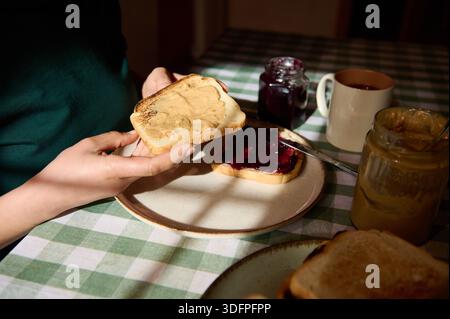 A warm, sunlit breakfast scene showing peanut butter being spread on bread with jelly nearby, on a checkered table with mugs and jars in the backgroun Stock Photo