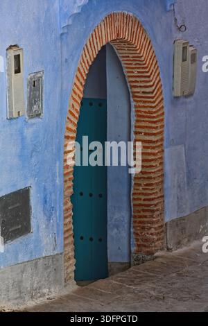 056 Azure, studded, wood door with red brick arch frame, set into the blue washed wall of a house on Calle Hassan I in the medina. Chefchaouen-Morocco Stock Photo