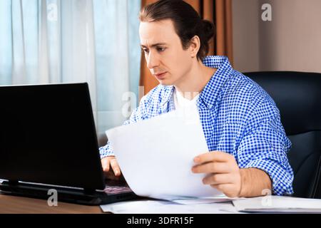 Serious man checking documents while working on a laptop. Concept of remote work, paying bills, and business administration in a home office. High qua Stock Photo