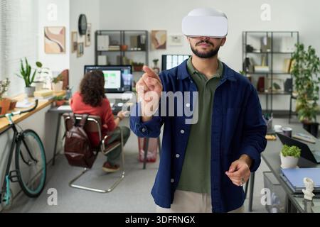 Young adult man wearing virtual reality headset interacting with invisible interface in modern office while colleague working at computer in background Stock Photo
