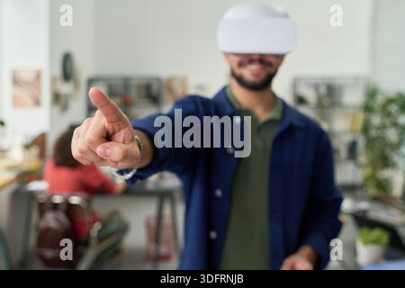 Young adult man wearing virtual reality headset interacting with digital interface, standing in modern workspace with outstretched arm and extended finger Stock Photo