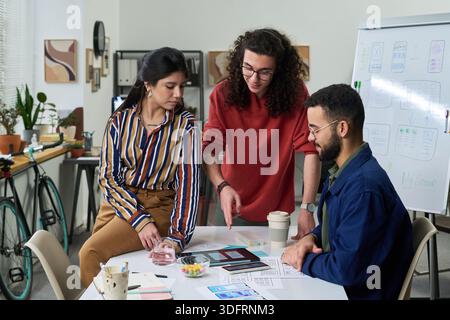 Multiethnic group of young adults collaborating on project, discussing digital interface designs at table with documents and tablet, creative workspace with brainstorming session Stock Photo