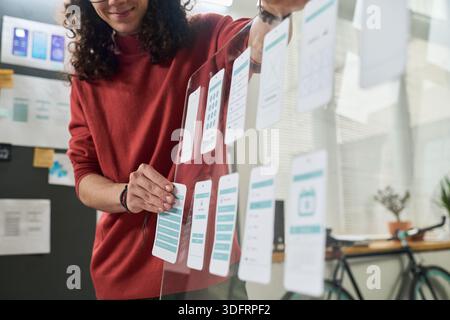 Young adult Caucasian man arranging mobile app interface mockups on transparent board, standing in modern office, focusing on user experience design process for digital project Stock Photo
