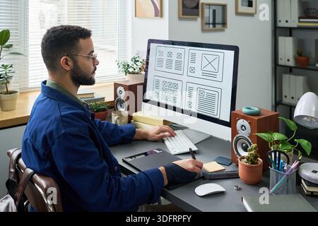 Young man sitting at desk working on computer with wireframe layouts on screen, using mouse and keyboard, surrounded by plants and office supplies in modern workspace Stock Photo