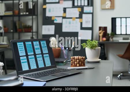 Modern office workspace featuring open laptop displaying mobile app interface design, desk accessories including potted plant and abacus, blurred background with shelving and wall charts Stock Photo