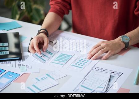 Unknown man working on mobile app interface design, arranging paper prototypes and wireframes on desk, using hands to organize layout and plan user experience Stock Photo