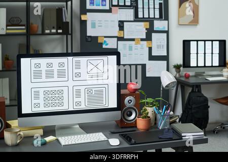 Modern workspace featuring large desktop monitor displaying wireframe layouts, digital tablet, speakers, potted plants, and organized desk setup with design sketches on wall in background Stock Photo