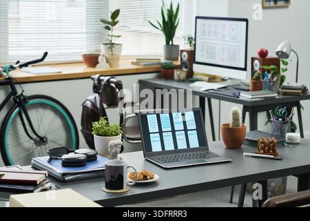 Modern workspace featuring open laptop displaying wireframe designs on desk, desktop monitor with interface layouts in background, bicycle near window, various plants and office supplies arranged Stock Photo