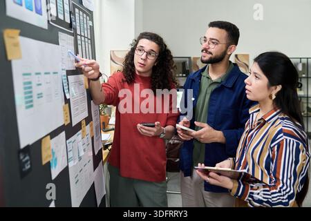 Multiethnic group of young adults collaborating on project, standing near board with charts and diagrams, holding digital tablets and smartphones, discussing strategy together Stock Photo