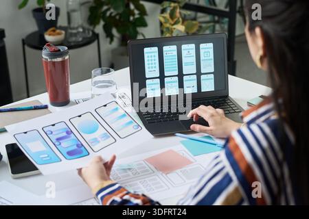 Young adult woman designing mobile app interface, holding wireframe sketches while working on laptop displaying smartphone screen layouts, sitting at desk with digital tablet and documents Stock Photo