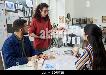 Multiethnic group of young adults collaborating on mobile app interface design, discussing wireframes and digital prototypes at creative office workspace, using tablet and printed layouts Stock Photo