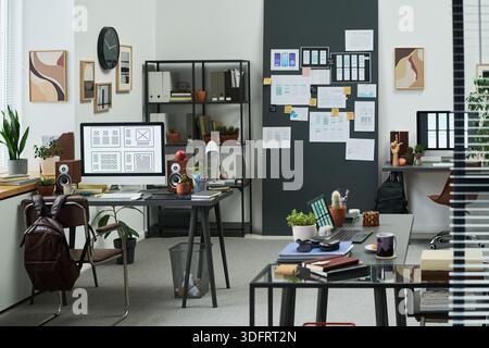 Modern office workspace showing empty desks with computers, digital devices, plants, and creative materials, wall covered with wireframes and notes, natural daylight illuminating scene Stock Photo