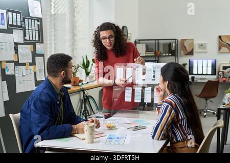 Young adult Caucasian man presenting mobile app wireframes to young adult multiethnic colleagues in modern office, team collaborating on user interface design project, discussing prototypes Stock Photo