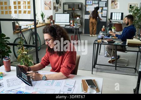 Caucasian young adult man using digital tablet while working on design project at desk in modern office, multiethnic young adult colleagues collaborating in background Stock Photo