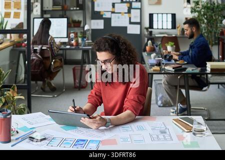 Young adult Caucasian man using digital tablet with stylus while working on design project at desk, two young adult colleagues collaborating in modern office background Stock Photo