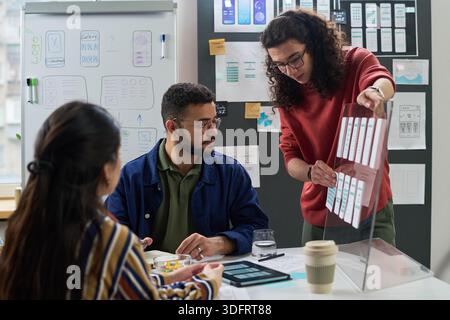 Multiethnic group of young adult coworkers collaborating on user interface design project, man presenting wireframe board while man and woman listening attentively Stock Photo