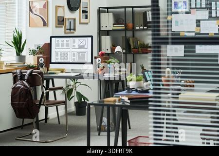 Modern office workspace featuring desktop computer with wireframe designs on screen, desk accessories, potted plants, books, shelves, and copy space for creative projects Stock Photo