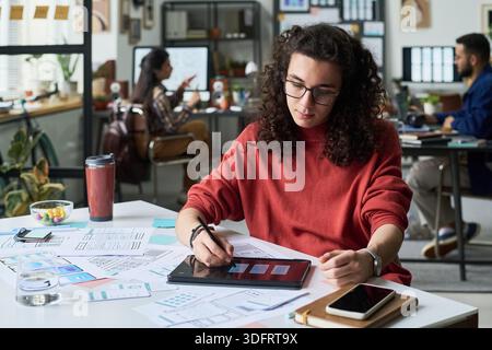 Young adult Caucasian man using digital tablet stylus while working on user interface design sketches at desk in modern office, colleagues collaborating in background, smartphone and coffee cup nearby Stock Photo