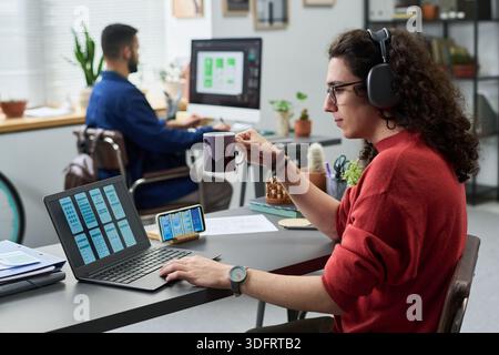 Young adult Caucasian man wearing headphones working on laptop with smartphone and holding coffee mug in modern office, male colleague with beard working at computer in background Stock Photo
