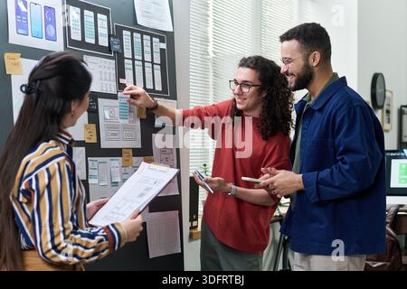 Multiethnic group of young adult men and woman collaborating on mobile app interface design, discussing wireframes and user experience concepts while standing near board with sketches Stock Photo