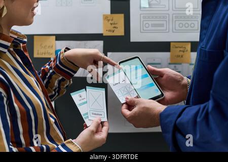 Young woman and man collaborating on mobile app interface design, holding smartphone and wireframe cards, discussing user experience in office setting Stock Photo