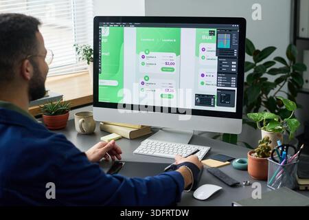 Man working on user interface design for mobile application on desktop computer, sitting at desk with digital pen and looking at large monitor displaying app screens Stock Photo