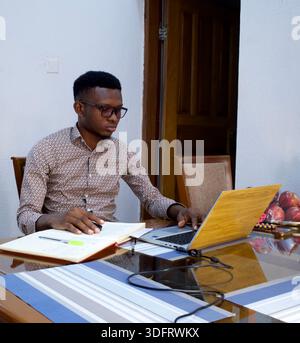 Young African man in glasses working on laptop and taking notes in open notebook at home desk in Nigeria. Stock Photo