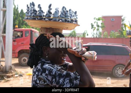 Young African woman street vendor balancing tray of bagged local nuts on head while handling money in Asaba, along Benin Expressway, Nigeria. Stock Photo