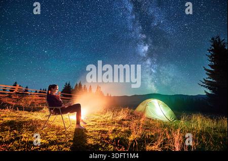 Hiker woman relaxes by campfire, illuminated by its warm glow, with tent nearby under stunning, star-filled sky. Milky Way stretches overhead, adding magical touch to serene mountain setting. Stock Photo