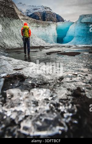 View of a lone explorer with a vibrant orange jacket trekking through the mesmerizing sapphire ice cave, Vatnajokull glacier, Iceland. Stock Photo