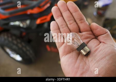 Close up view showing hand holding small automotive light bulb with blurry orange ATV vehicle background capturing preparation for repair maintenance Stock Photo