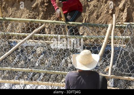 Two workers focused on construction project building sturdy retaining wall with wire mesh gabion basket filled with heavy rock stone near dirt mound Stock Photo