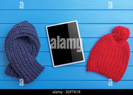 A digital tablet with a black screen placed between a navy blue knitted scarf and a red pom-pom beanie on a blue wooden surface. Stock Photo