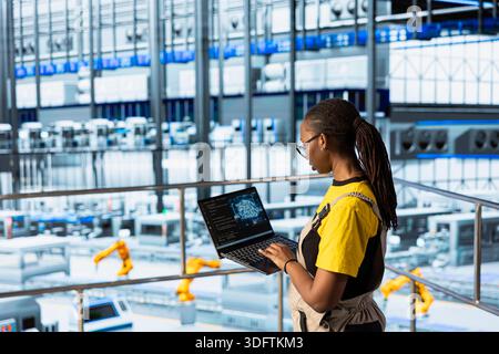Smart factory female technician implements automated workflows using artificial intelligence tech, computerizing operations. African american programmer integrates sensors into production using AI Stock Photo