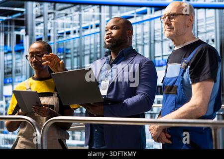 Manager using laptop to brief diverse team of solar panel factory workers on elevated platform. Supervisor discussing automated workflow plans with technician and female engineer in industrial plant Stock Photo