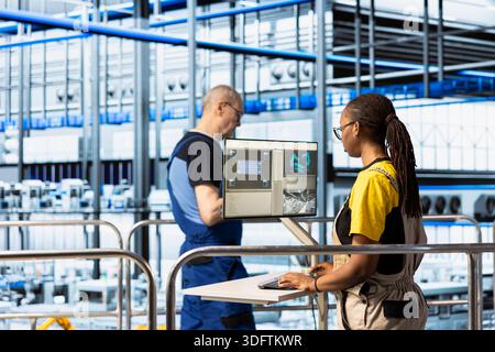 Factory worker operating control panel on elevated platform overlooking automated production line. Female engineer managing industrial machinery workflow using digital interface Stock Photo