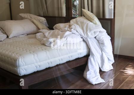 White messy bed with unmade duvet and white pillow on wooden bed frame in bedroom showing lazy or relaxed morning atmosphere with sunlight shining on Stock Photo