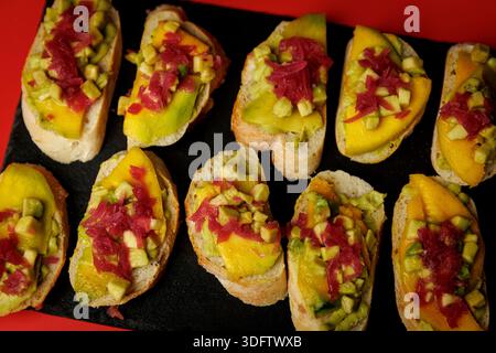 Top view of small bruschetta slices topped with mango, avocado and pickled red onion on a black slate tray with red background. Colorful vegetarian fi Stock Photo