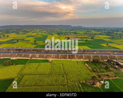 Aerial view of vibrant green and yellow fields intersected by a modern highway under a soft sky, Alwar, Rajasthan, India. Stock Photo