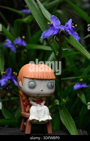 Funko Pop action figure of haunted doll Annabelle sitting on chair from The Conjuring horror films. Creepy doll in dark garden among blue flowers. Stock Photo