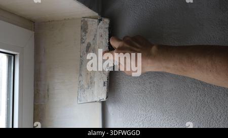 A hand with a wide trowel is leveling the surface of the slope of a small window from inside the room, smoothing the plaster on the window sill Stock Photo
