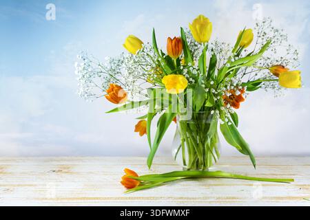 Cheerful bouquet of flowers with orange and yellow tulips and some gypsophila on a light wooden table in front of a blue sky, Easter decoration, sprin Stock Photo