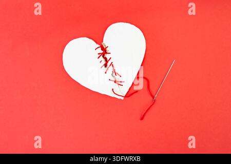 Broken cardboard heart sewed together with a needle and red thread on red background, marriage  concept Stock Photo