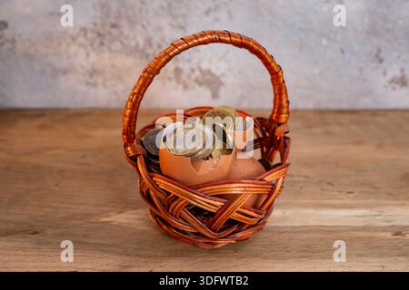 Broken egg shells filled with coins in a woven willow basket, on wooden desk, all the eggs in one basket,  investment metaphor Stock Photo