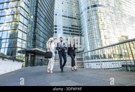 Diverse business professionals walking, communicating, and laughing while moving together in a modern financial district with glass office buildings r Stock Photo