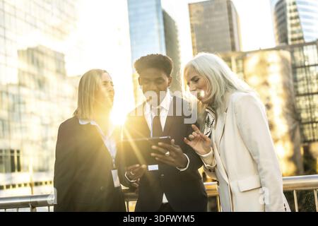 Diverse multi generation business team outdoors at sunset, collaborating over a digital tablet amid urban skyscrapers teamwork, technology and leaders Stock Photo