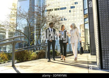 Three diverse business professionals, a woman, a man, and a senior woman, walking and actively discussing outdoors in a modern city setting with glass Stock Photo