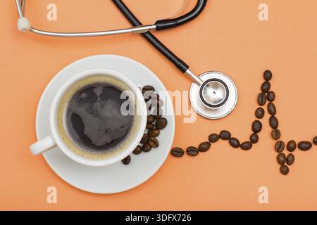 White cup of coffee on saucer with coffee beans folded in the form of a cardiogram and phonendoscope on peach colored background. Top view Stock Photo