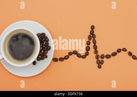 White porcelain cup of coffee on saucer with coffee beans folded in the form of a cardiogram on peach colored background. Top view Stock Photo