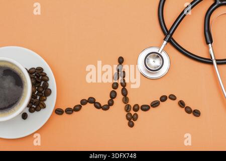 White porcelain cup of coffee on saucer with coffee beans folded in the form of a cardiogram and phonendoscope on peach colored background. Top view Stock Photo